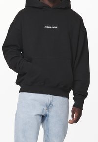 Svart munkjacka med huva och framficka, ribbade muddar och fåll, med vit "PEGADOR"-logotyp på bröstet. Tillverkad av bomull, avslappnad passform.