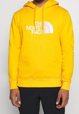 Mies, joka käyttää kirkkaan keltaista hupparia, jossa on valkoinen "The North Face" -logo, mustat housut, seisoo yksivärisen vaalean taustan edessä.