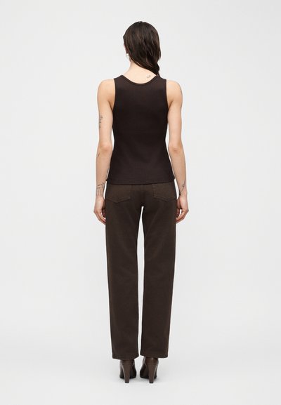 Γυναίκα με μακριά σκούρα μαλλιά, φορώντας μαύρο αμάνικο τοπ, σκούρο καφέ παντελόνι και καφέ παπούτσια με τακούνι, όρθια με την πλάτη προς το μέρος του θεατή σε απλό φόντο.