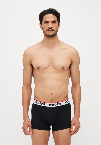 Sorte boxer shorts med en hvid elastisk talje med rød "MOSCHINO" tekst. Modellen står vendt fremad og fremviser pasformen.