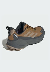 Brown athletic shoe with a textured fabric upper, gray rubber sole, and three light gray stripes. Features reinforced toe and heel areas.