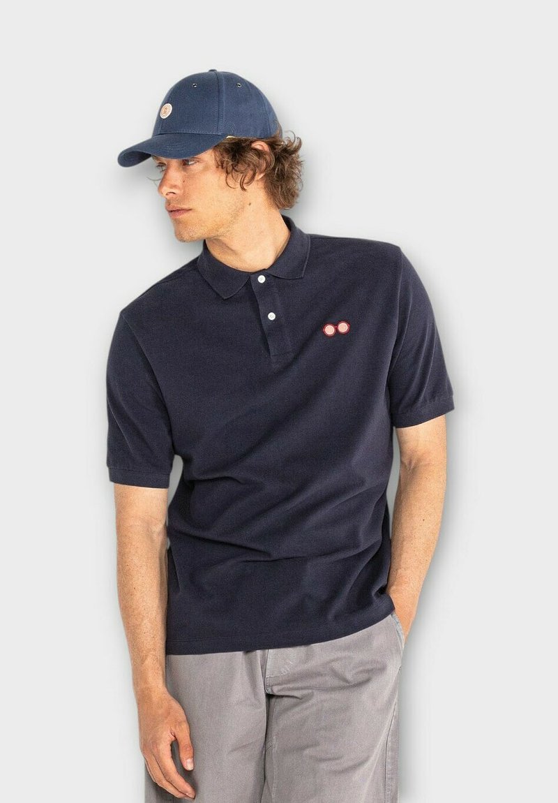 Marineblauw poloshirt met korte mouwen, drie knopen bij de hals, en drie rode cirkelvormige accenten op de borst. Gecombineerd met een bijpassende pet.