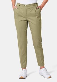 Hellolivgrüne, taillierte Hose mit zwei vorderen Taschen und einem Reißverschluss, die durch klare Linien und eine glatte Textur besticht, kombiniert mit weißen Sneakers.