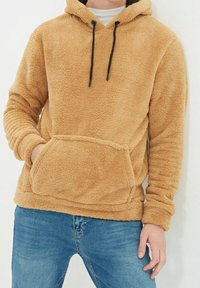 Beżowa bluza polarowa o fakturowanej powierzchni, wyposażona w przednią kieszeń, regulowany kaptur ze sznurkiem oraz ściągacze na rękawach. W zestawie z niebieskimi dżinsami.