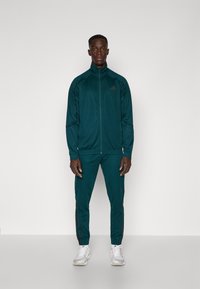 Turkizna atletska trenirka z jopičem na zadrgo in tesno prilegajočimi hlačami. Črne kontrastne črte ob straneh. Lahka, gladka tkanina.