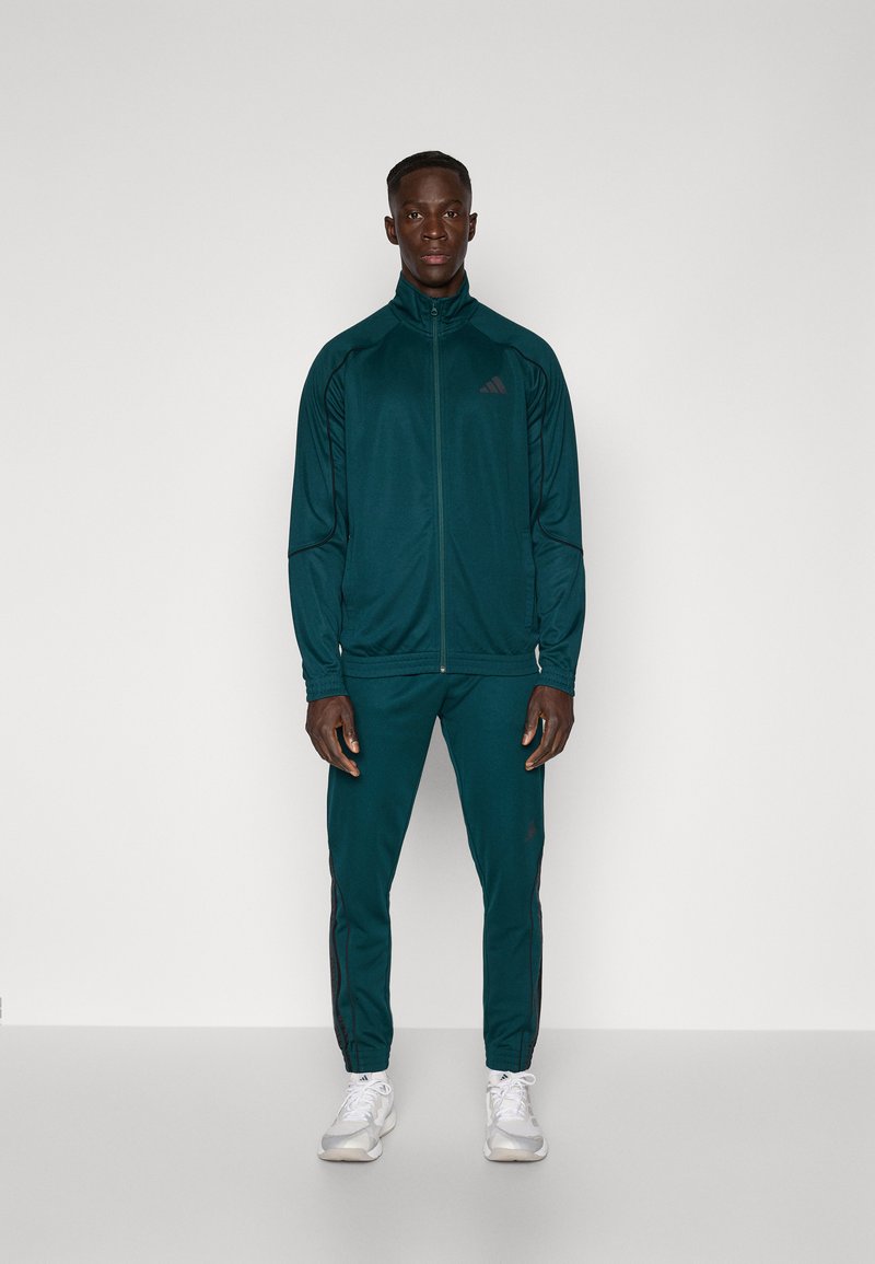 Turkizna atletska trenirka z jopičem na zadrgo in tesno prilegajočimi hlačami. Črne kontrastne črte ob straneh. Lahka, gladka tkanina.