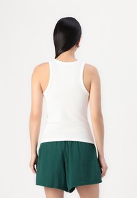 Weißes, geripptes Tanktop mit rundem Ausschnitt, kombiniert mit grünen, strukturierten, weit geschnittenen Shorts. Von hinten betrachtet.