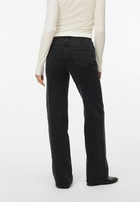 Personne portant un jean noir taille haute à jambes droites avec poches arrière et un haut blanc ajusté à manches longues, debout sur un fond blanc.