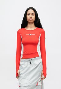 Czerwona bluzka z długim rękawem, z białymi panelami po bokach i logo. Materiał jest gładki, dopasowany, zestawiona z jasnoszarymi szortami z sznurkiem.