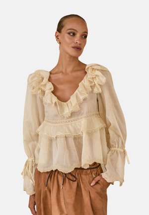 Kobieta w kremowej, falbaniastej bluzce z długim rękawem i koronkowym wykończeniem oraz brązowych spodniach, ręka w kieszeni, patrząca na bok.