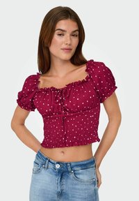 Frau in einem kurzärmligen, burgunderfarbenen Crop-Top mit weißen Punkten und Jeans, die mit den Händen hinter dem Rücken steht.