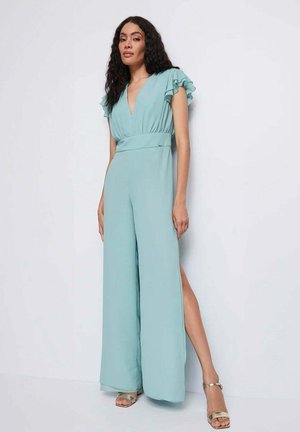 Tuta azzurro chiaro con maniche corte arricciate, scollo a V profondo, vita con cintura, silhouette a gamba larga e spacchi laterali, abbinata a sandali metallici.