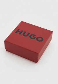 Κόκκινο χαρτονένιο κουτί με ματ φινίρισμα, διακοσμημένο με έντονο μαύρο κείμενο «HUGO» στην κορυφή, τετράγωνο σχήμα και καθαρές άκρες.
