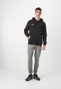 Zwarte hoodie met een voorzak en wit logo, gecombineerd met grijze joggingbroek en zwarte sport schoenen. Eenvoudig ontwerp, comfortabele pasvorm.