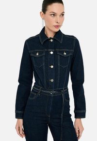 Giacca in denim scuro con silhouette strutturata, bottoni dorati, due tasche sul petto e un cinturino decorato con borchie. Presenta maniche lunghe.