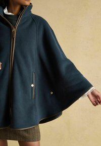 Námornícky fleecový pončo s vysokým golierom, zlatým zipsom, prednými vreckami a kontrastnými ozdobami. Má široké rukávy a textúrovaný lem.