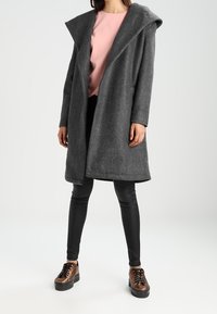 Abrigo de lana gris con una gran capucha, frente abierto y mangas largas, combinado con leggins negros y zapatos de cordones marrones.