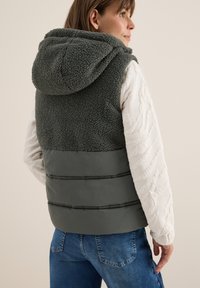 Faux furväst i mörkgrön med en huva; har en kviltad tygpanel i grått, detaljerad sömnad och en figursydd design.