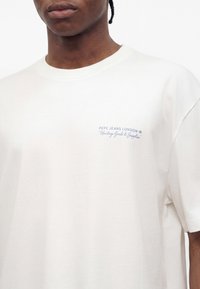 Άτομο που φοράει λευκό T-shirt με το κείμενο «PEPE JEANS LONDON Heritage Goods & Supplies» στο αριστερό στήθος.