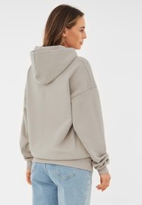 Hanorac gri deschis cu glugă, cu croială lejeră, umeri lăsați și manșete cu model canalé, asortat cu blugi albaștri deschiși. Textură netedă.