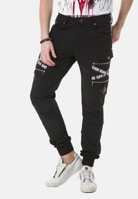 Schwarze Cargo-Hosen mit Reißverschlusstaschen, elastischen Bündchen und Druckakzenten. Hergestellt aus einem Stoff mit glatter Oberfläche. Weiße Sneakers runden den Look ab.