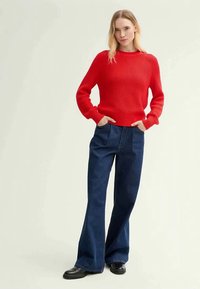 Sweater de malha canelada vermelha com mangas longas, combinado com jeans de boca de sino azul escuro e botas pretas, criando um visual casual.