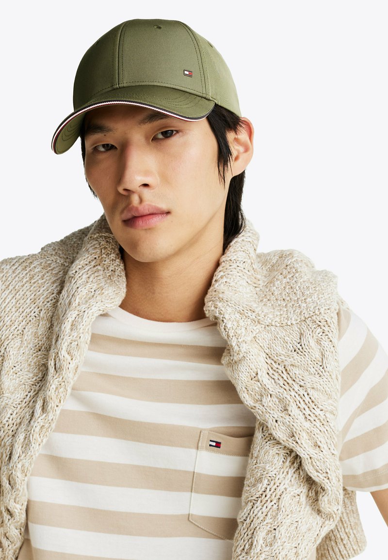 Cappellino da baseball verde oliva con dettagli di cucitura a contrasto rossi e bianchi, indossato con una camicia leggera a righe beige e crema sotto un gilet di maglia.