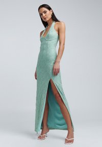 Robe vert clair à col licou avec une finition texturée, présentant une fente haute et une silhouette ajustée, associée à des sandales à talons blanches.