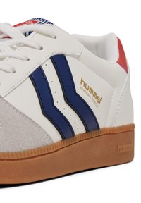 White sneaker with suede accents, blue and black stripes, and red heel. Features white laces and a gum outsole. "Hummel" logo in gold.