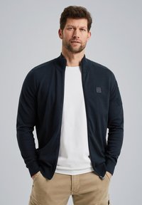 Black zip-up cardigan made of soft fabric, features side pockets and a small logo patch on the chest. Worn over a white t-shirt.