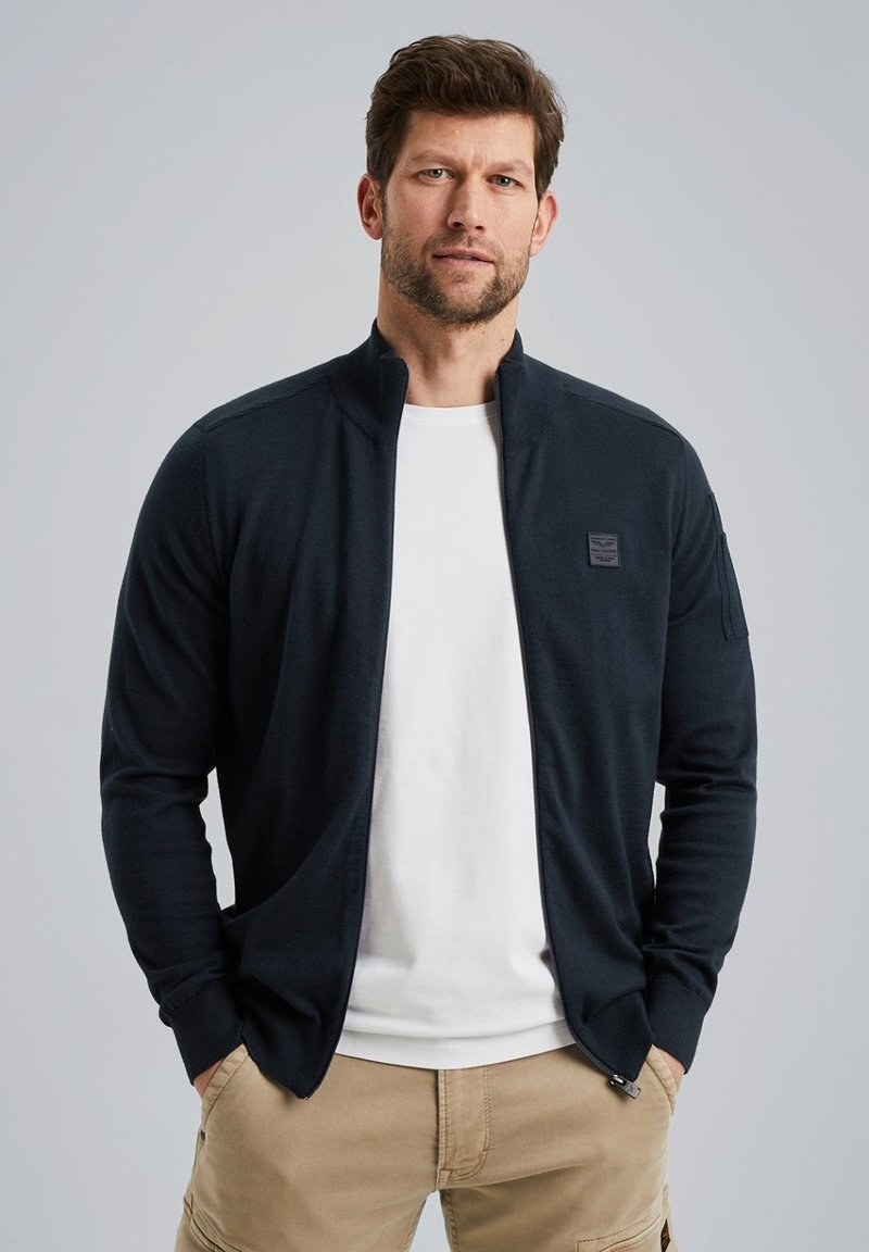 Black zip-up cardigan made of soft fabric, features side pockets and a small logo patch on the chest. Worn over a white t-shirt.