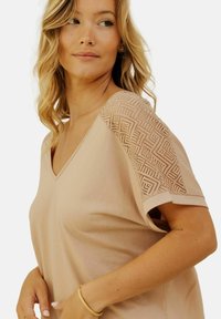 Top en V beige clair avec des détails en dentelle géométrique sur l'épaule. Tissu doux, coupe décontractée et manches courtes retournées.