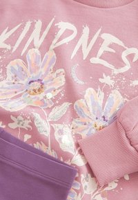 Ljusrosa sweatshirt med blommotiv prydd med paljetter, ordet "KINDNESS" i fet vit text och en kontrasterande lila ficka.