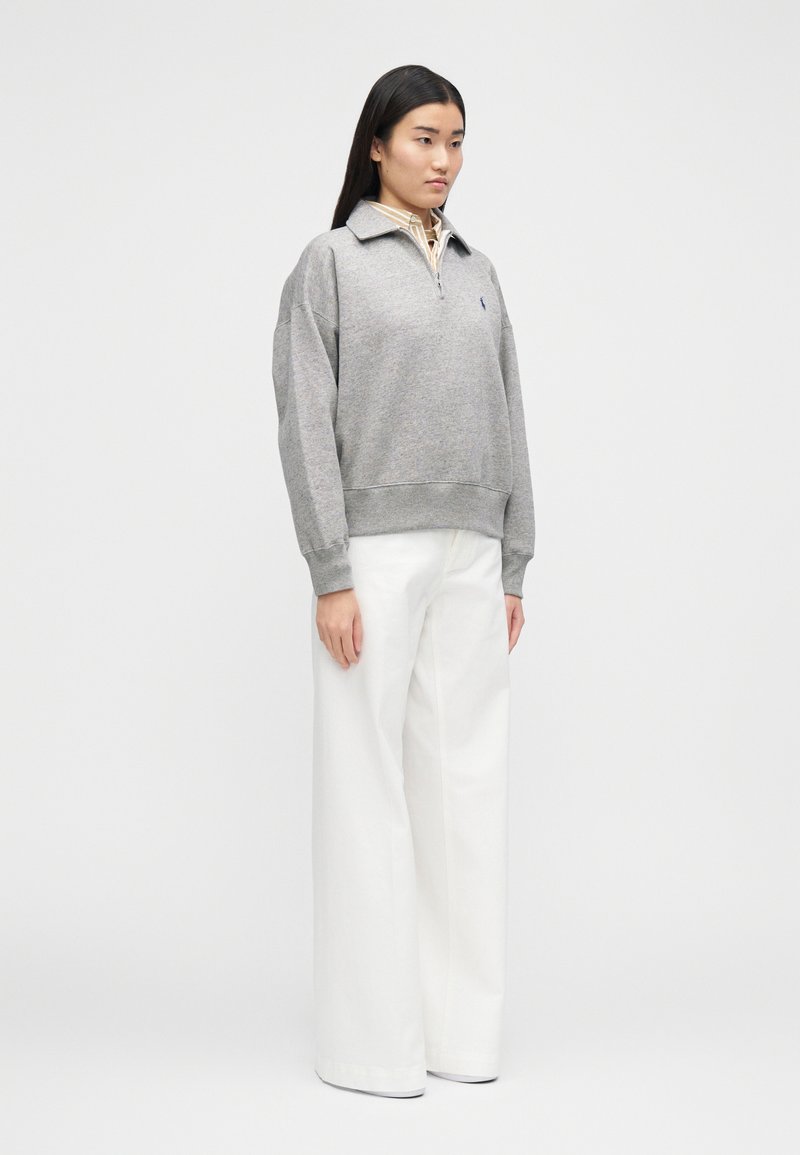 Pull gris à col avec fermeture éclair, porté par-dessus une chemise rayée claire, associé à un pantalon blanc à jambes larges. Design simple et décontracté.