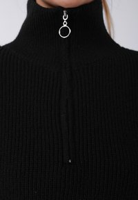 Pull noir à col montant côtelé avec une fermeture éclair argentée munie d'un tirette rond. La texture est tricotée, mettant en avant chaleur et confort.