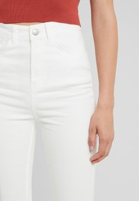 Kvinna som bär högmidjade vita jeans med knäppning och en croppad rustfärgad topp, armen avslappnad vid sidan.