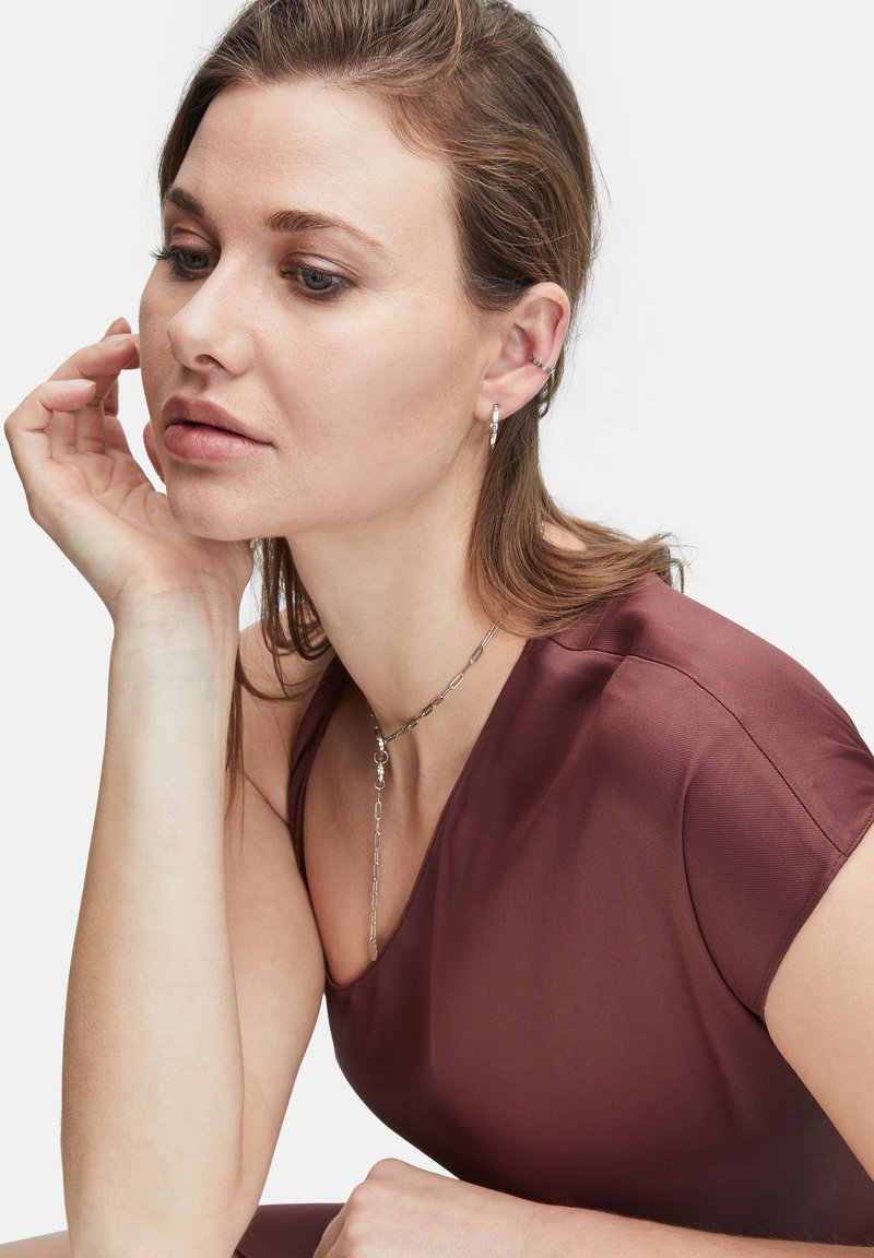 Collana in argento con maglie allungate, caratterizzata da un ciondolo centrale. Indossata con un top corto, maniche corte di colore bordeaux, con una texture liscia.