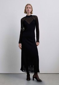 Falda larga negra acanalada con dobladillo con flecos combinada con un top de manga larga translúcido con rayas. La modelo sostiene un pequeño bolso negro y lleva sandalias con tacón.