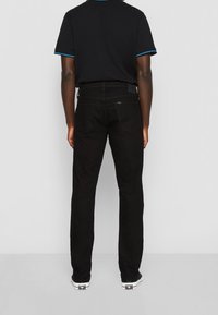 Personne portant un jean noir, un polo noir avec des bordures bleues aux manches et des baskets blanches, debout de dos devant un fond uni.