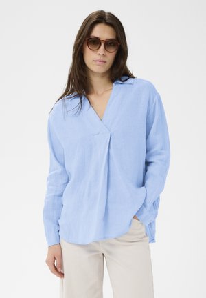 Nő teknősbéka mintás napszemüvegben, világoskék, hosszú ujjú, V-nyakú blúzban és bézs nadrágban, egyik kezét a zsebében tartva pózol.