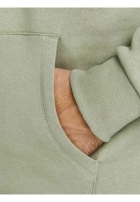 Main reposant dans la poche d'un sweat-shirt vert clair avec un poignet côtelé visible.