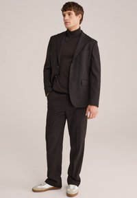 Mann in einem dunkelbraunen Anzug mit einem Rollkragenpullover und weißen Sneakers, der vor einem neutralen Hintergrund steht.