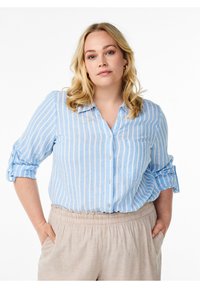Camicia azzurra e bianca a righe con maniche arrotolate e taschino sul petto, abbinata a pantaloni beige con vita elastica.