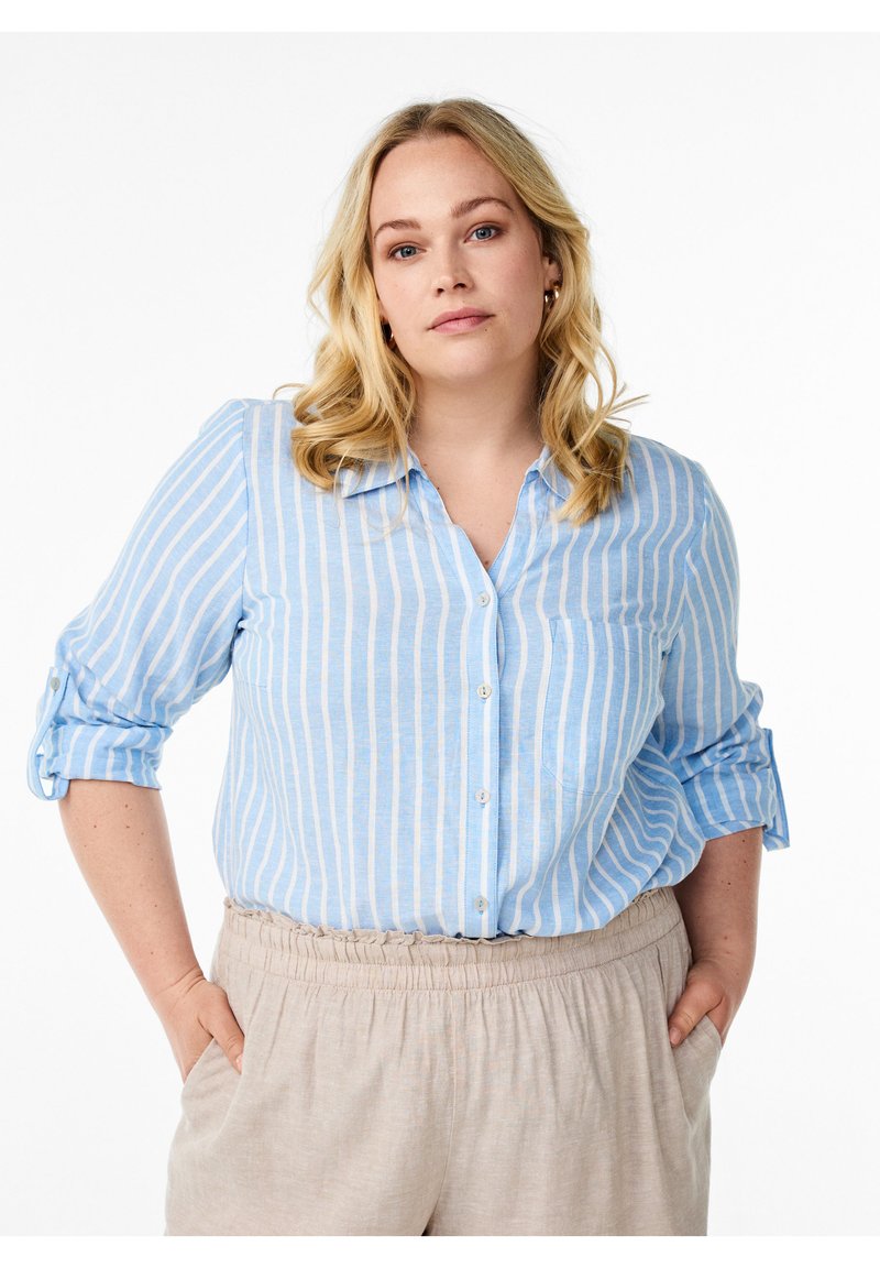 Camicia azzurra e bianca a righe con maniche arrotolate e taschino sul petto, abbinata a pantaloni beige con vita elastica.