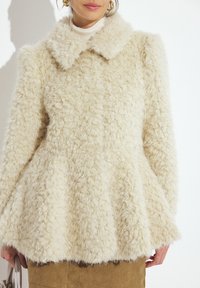 Cremefarvet, fluffy faux fur frakke med en bred krave, tekstureret overflade og peplum-silhuet. Har lange ærmer og en skjult lukning.