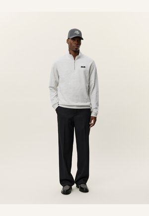 Gray zip-up fleece pullover with a black logo patch, paired with dark, loose-fitting trousers and a gray cap with embroidered detail.