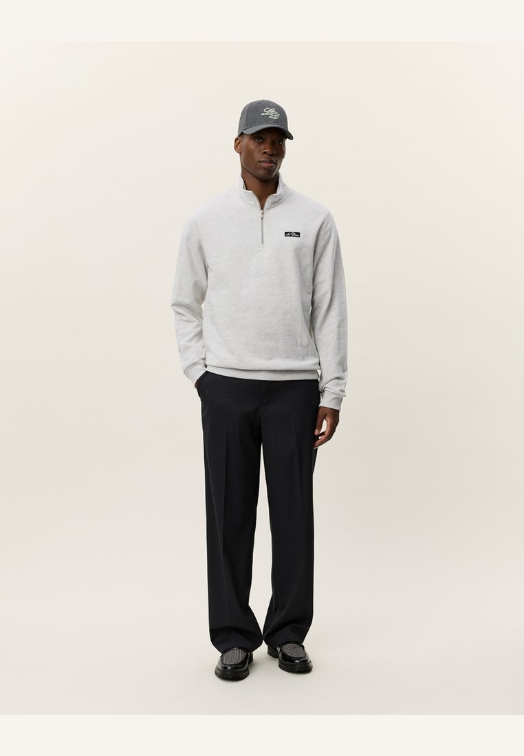 Grå zip-up fleece-pullover med et sort logo-patch, parret med mørke, løstsiddende bukser og en grå cap med broderet detalje.