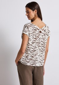 Camicia a maniche corte color crema con strisce zebra marroni, caratterizzata da un design con schiena a V e un piccolo dettaglio a taglio sul collo.