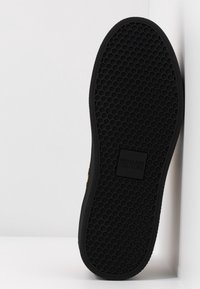 Semelle de chaussure noire avec un motif hexagonal et un patch logo rectangulaire portant "Versace Jeans Couture" au centre.