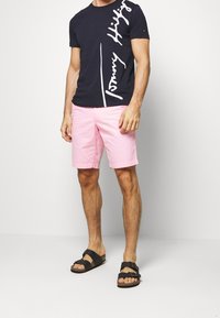 T-shirt in cotone navy con logo bianco, abbinato a pantaloni corti rosa chiaro in un tessuto liscio, e sandali neri con cinturini regolabili.